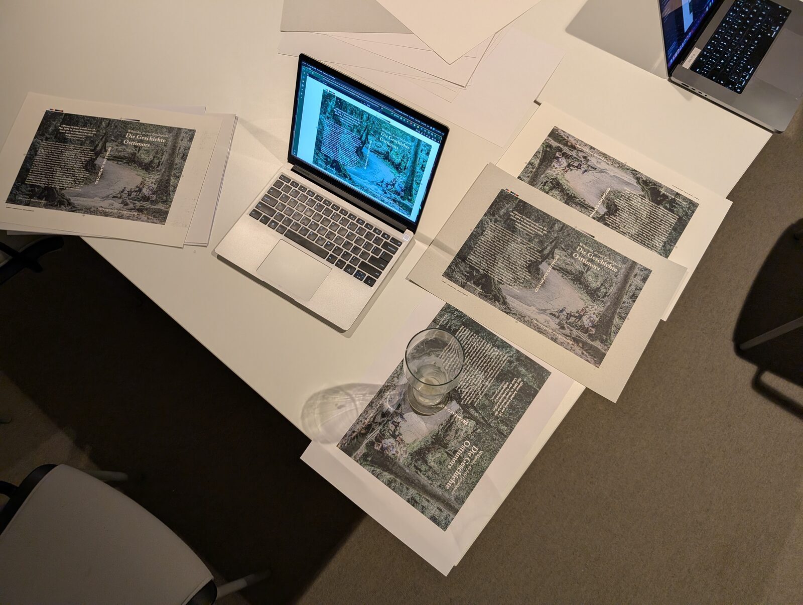 multiple print samples of the same cover design scattered on a table, two open laptops